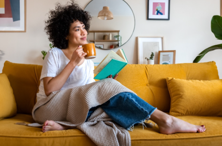 Femmes lisant un livre et dégustant une tasse de thé sur son canapé avec une couverture sur ses genoux.